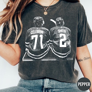 Könnte beinhalten: Dunkelgraues T-Shirt mit einer Schwarz-Weiß-Grafik von zwei Eishockeyspielern von hinten, die Trikots mit den Namen "CELEBRINI" und "SMITH" und den Nummern "71" und "2" tragen. Das T-Shirt hat einen Vintage-Look.