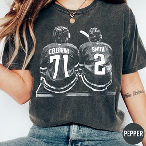 Könnte beinhalten: Dunkelgraues T-Shirt mit einer Schwarz-Weiß-Grafik von zwei Hockeyspielern. Das Trikot links hat den Namen "CELEBRINI" und die Nummer "71". Das Trikot rechts hat den Namen "SMITH" und die Nummer "2".