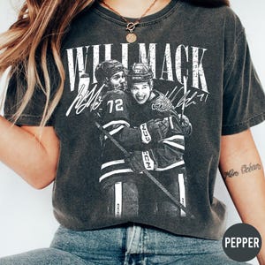 Könnte beinhalten: Dunkelgraues T-Shirt mit einer Grafik von zwei sich umarmenden Eishockeyspielern, darüber der Text "WILL MACK" und ihre Unterschriften. Die Spieler tragen Hockeytrikots und Helme und halten einen Hockeyschläger.