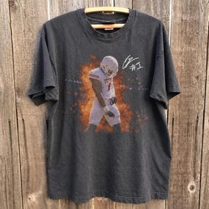 Op de afbeelding: Een antracietgrijs T-shirt met korte mouwen en een afbeelding van een footballspeler in een wit uniform met de tekst TEXAS en het nummer 1, omringd door oranje vlammen. Een witte handtekening is rechts boven de speler gedrukt.