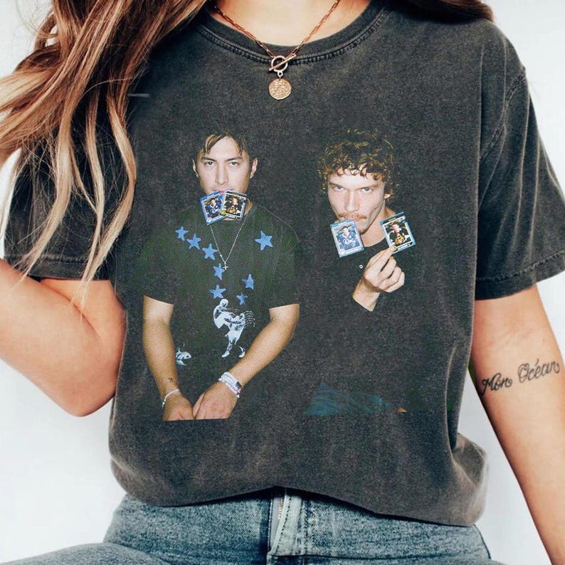 May include: Dark gray t-shirt featuring a graphic of two men holding cards. The shirt has a vintage, worn look. One man's shirt has blue star and figure graphics. The other man's shirt is plain. The shirt is paired with blue jeans.