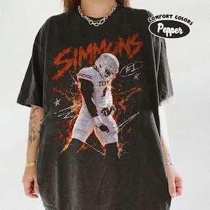 Op de afbeelding: Een antracietgrijs t-shirt met een afbeelding van een footballspeler in een wit uniform met de naam SIMMONS en het nummer 1. Het ontwerp bevat oranje en witte steraccenten, de tekst #1 en een Comfort Colors Pepper label.