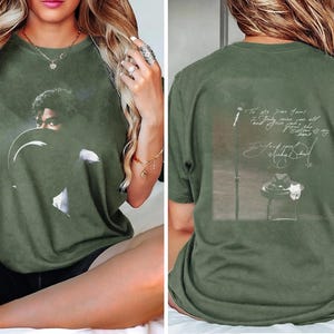 May include: Olive green t-shirt featuring a portrait of a person with a hat on the front. The back of the shirt has a graphic with text and a signature. The shirt is a relaxed fit.