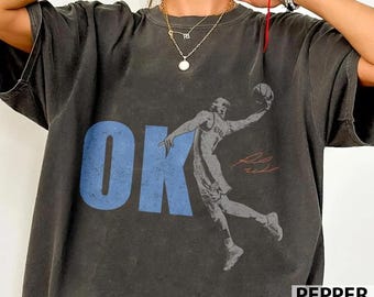 May include: A charcoal grey short-sleeve t-shirt featuring a large blue graphic of the letters OK and a black and white illustration of a basketball player in mid-air. The design includes a red signature graphic next to the player illustration.