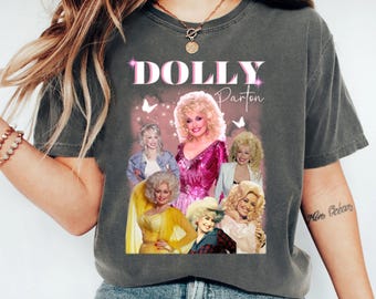 May include: A charcoal gray t-shirt featuring a graphic print of Dolly Parton in various outfits and poses. The text "DOLLY" appears in large white and pink letters above the images, with "Parton" written in a cursive script to the right.