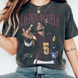 May include: A charcoal grey t-shirt featuring a graphic print of Jayden Daniels in a maroon and gold football jersey with the number 5. The graphic shows the player holding a beer bottle, smoking, and gesturing with a middle finger, with the text JAYDEN DANIELS and a signature.