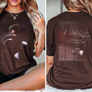 May include: Brown t-shirt featuring a portrait of a musician. The front of the shirt displays a close-up image of the musician. The back of the shirt has a graphic of a microphone, a hat, and text that reads "To my dear fans..."