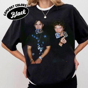 May include: Black t-shirt with a graphic of two people holding cards. The shirt has a crew neck and short sleeves. The text "COMFORT COLORS Black" is printed on the upper left side of the shirt.