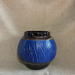 May include: A round ceramic vase featuring a textured, crackled cobalt blue center band contrasted by smooth, dark brown glazed sections at the top and bottom. A thin, gold-colored line follows a crack along the upper rim of the vessel.