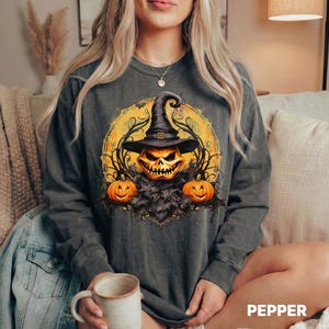 Puede incluir: Una sudadera gris carbón de manga larga con un estampado gráfico de una calabaza grande y sonriente con un sombrero de bruja negro, flanqueada por dos calabazas más pequeñas sobre un fondo circular amarillo con enredaderas.