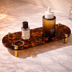 Puede incluir: Una bandeja decorativa con un patrón de carey y patas doradas exhibe un frasco de perfume, un producto para el cuidado de la piel y joyas doradas. La bandeja está sobre una superficie de mármol.