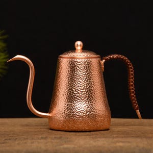 May include: Copper-colored gooseneck kettle with a textured surface and a woven brown handle. The kettle has a long, curved spout and a small lid with a round knob. The kettle is designed for pour-over coffee brewing.