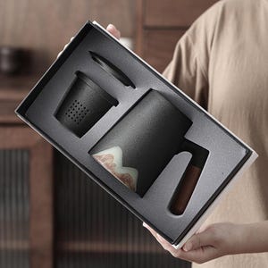 May include: A black ceramic tea mug set with a wooden handle, a tea infuser, and a lid, presented in a gift box. The mug features a mountain design in shades of white, brown, and green. The set is designed for tea lovers.