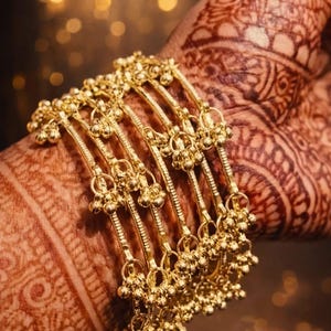 Op de afbeelding: Een close-up toont een set gouden armbanden. Elke armband heeft een delicaat ontwerp met kleine, bungelende kralen. De armbanden worden getoond op een hand versierd met ingewikkelde henna-patronen, wat een mooi contrast creëert.