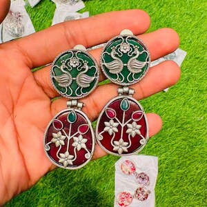 May include: Silver earrings with green and red enamel detailing. The top pair features a green background with peacock motifs. The lower pair has a red background with floral designs. Additional earrings are visible in the background.