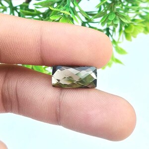 Può includere: Una gemma rettangolare sfaccettata con una tonalità fumosa, marrone-verde. La gemma è tenuta tra due dita, mostrando i suoi tagli geometrici e la superficie lucida. Il colore della gemma varia da chiaro a scuro, con una leggera trasparenza.