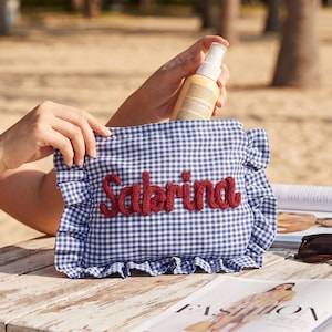 May include: A blue and white gingham fabric pouch featuring a ruffled border and the name Sabrina embroidered in red yarn across the centre. The rectangular pouch has a soft, textured appearance and is designed with a gathered fabric edge.