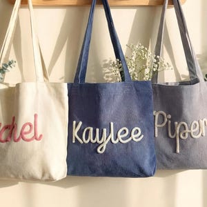 Może przedstawiać: Trzy torby typu tote z sztruksu wiszące na drewnianym wieszaku. Kremowa torba ma napis Rachel w kolorze różowym, niebieska torba ma napis Kaylee w kolorze białym, a szara torba ma napis Piper w kolorze białym. Każda torba ma dwa paski na ramię.