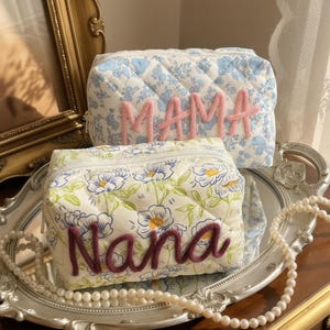 May include: Two quilted cosmetic bags. One is white with a floral pattern and the word "Nana" in purple. The other is blue with a floral pattern and the word "Mama" in pink. Both bags have a zipper closure.