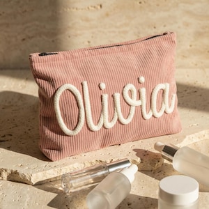 May include: A pink corduroy cosmetic bag with the name "Olivia" embroidered in white script. The bag is next to several clear glass bottles and jars, likely for skincare products. The items are arranged on a light-colored stone surface.