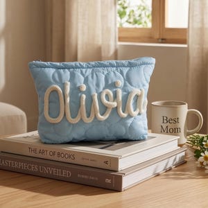 Op de afbeelding: Een lichtblauwe gewatteerde make-uptas met de naam "Olivia" in witte letters. De tas ligt op twee boeken, naast een mok met de tekst "Best Mom" en een klein boeket witte bloemen.