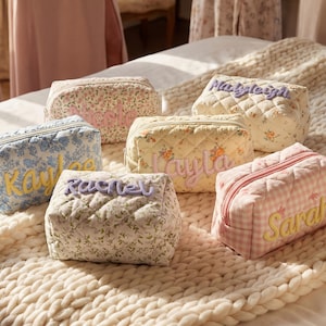 May include: A collection of personalized cosmetic bags in various floral patterns and colors. Each bag features a different name embroidered in a contrasting color. The bags are arranged on a cream-colored, textured blanket.