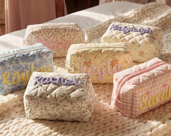 May include: A collection of personalized cosmetic bags in various floral patterns and colors. Each bag features a different name embroidered in a contrasting color. The bags are arranged on a cream-colored, textured blanket.