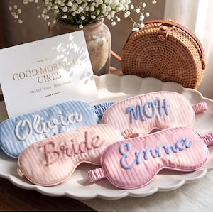 May include: Four satin sleep masks with striped textures and embroidered names. One light blue mask reads Olivia, one pink mask reads Bride, one pink mask reads MOH, and one pink mask reads Emma. Each mask features a matching elasticated strap.