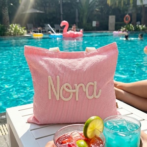 May include: A red and white striped tote bag with the name "Nora" embroidered in cream-colored thread. The bag has cream-colored handles and is sitting on a white table next to two cocktails. The background is a swimming pool.
