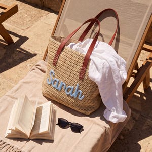 May include: A woven straw tote bag featuring brown leather handles and the name Sarah embroidered in light blue script. The bag is accompanied by a white linen shirt, an open book with white pages, and black sunglasses resting on a beige towel.