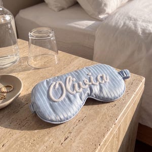 May include: A light blue sleep mask featuring a subtle vertical striped pattern and the name Olivia embroidered in white script lettering across the center. The mask includes an elasticated strap for a secure fit around the head.