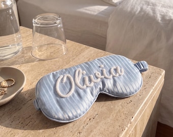 May include: A light blue sleep mask featuring a subtle vertical striped pattern and the name Olivia embroidered in white script lettering across the center. The mask includes a matching elastic strap for a secure fit around the head.