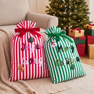 May include: Two fabric gift sacks with vertical stripes. The left sack is red and white with a red ribbon and the text "Joyful Giving" in raised letters, plus a reindeer and tree icon. The right sack is green and white with a green ribbon and the text "Merry Christmas" in raised letters, plus a Santa icon.