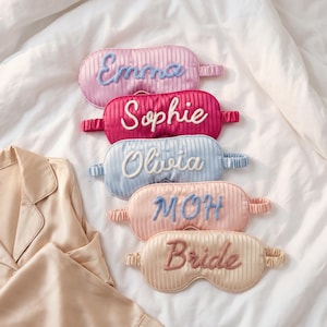 May include: Five oval-shaped sleep masks with a vertical striped texture and elastic headbands. The masks feature embroidered names and titles: Emma in blue, Sophie in white, Olivia in white, MOH in blue, and Bride in dark pink thread.