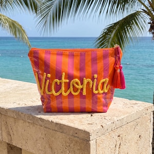 May include: A rectangular cosmetic pouch featuring vertical orange and pink stripes with a glossy finish. The name Victoria is embroidered in yellow cursive script on the front. A bright orange and pink tassel hangs from the side zipper pull.