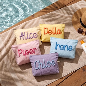 May include: Five colorful cosmetic bags with embroidered names: Alice, Della, Piper, Chloe, and Irene. The bags are in shades of yellow, pink, blue, and purple, arranged on a towel near a pool. A straw hat and sunglasses are also visible.