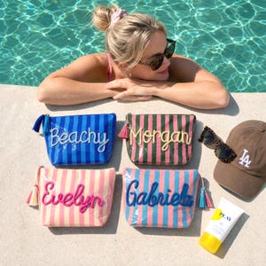 May include: Four rectangular zippered pouches with vertical stripes and personalized names in cursive script. The pouches feature blue, green, pink, and light blue color schemes with matching tassels attached to the side zippers.