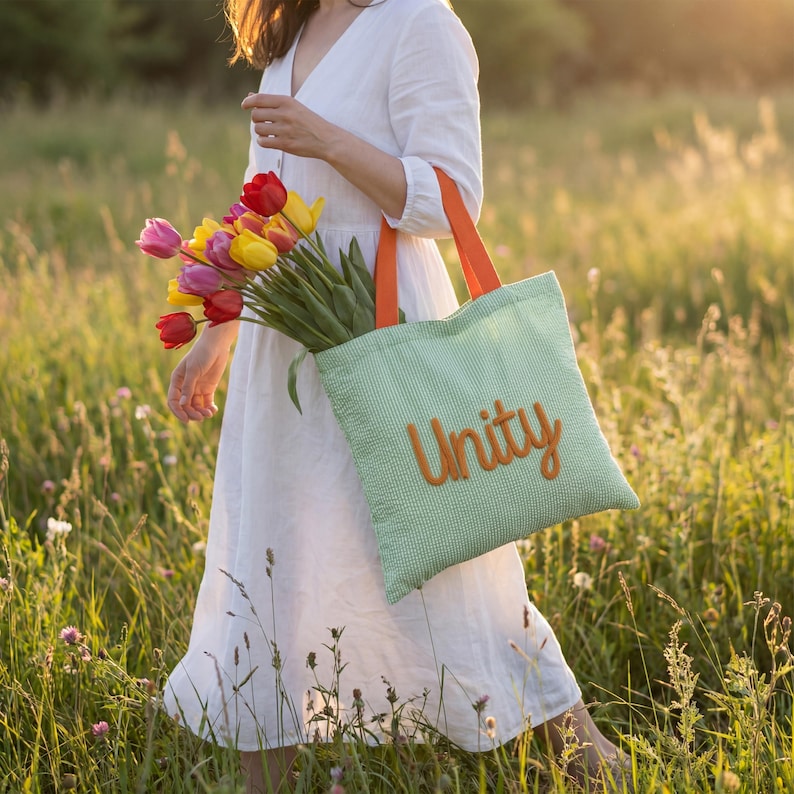 May include: A green tote bag with orange handles and the word "Unity" embroidered in orange. The bag is filled with a bouquet of colorful tulips. The person is wearing a white dress and walking through a field of tall grass.