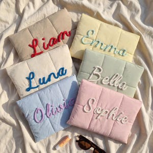 May include: Six rectangular quilted pouches in beige, yellow, cream, light green, light blue, and pink. Each pouch features a name embroidered in raised yarn: Liam in red, Emma in light green, Luna in blue, Bella in white, Olivia in purple, and Sophie in white.
