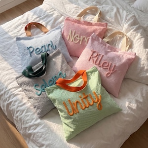 May include: Five striped tote bags in various colors, including blue, pink, and green, with embroidered names: "Pearl," "Nora," "Riley," "Selena," and "Unity." Each bag has a contrasting handle color, such as orange or green, and a matching embroidered name.