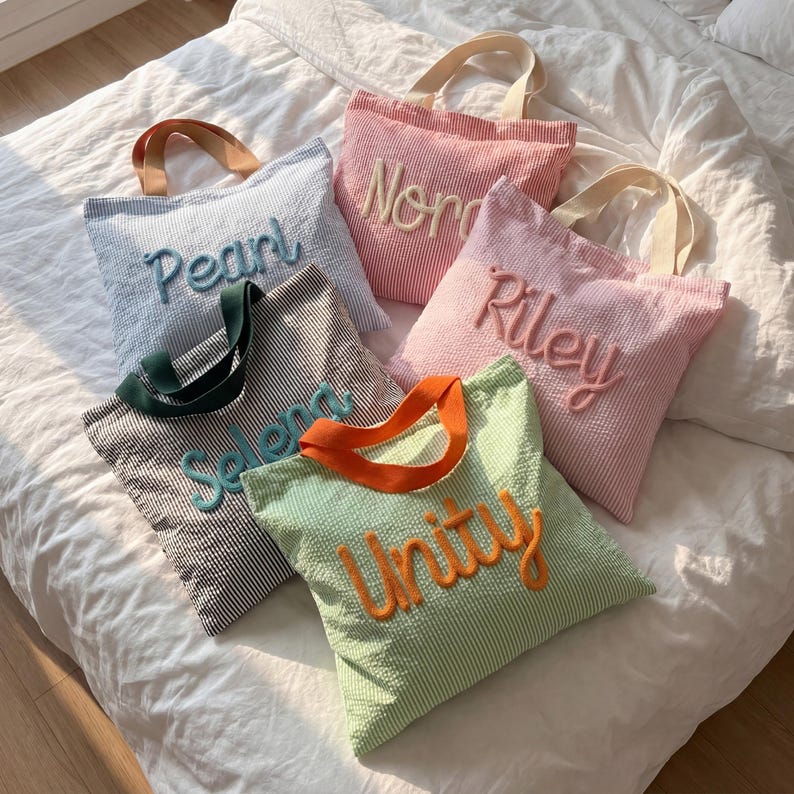 May include: Five striped tote bags in various colors, including blue, pink, and green, with embroidered names: "Pearl," "Nora," "Riley," "Selena," and "Unity." Each bag has a contrasting handle color, such as orange or green, and a matching embroidered name.