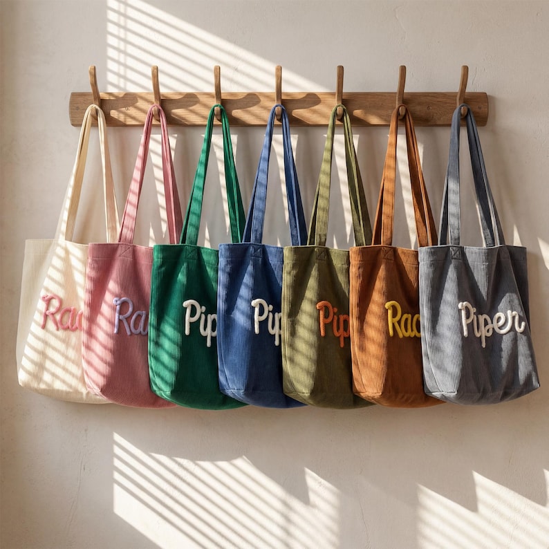May include: A row of seven corduroy tote bags in cream, pink, green, blue, olive, brown, and gray, each featuring a contrasting embroidered name in cursive script. The bags are hanging from a wooden rack with seven hooks against a plain wall.