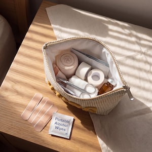 May include: A beige and white striped zippered pouch filled with medical supplies including a rolled bandage, adhesive tape, metal scissors, and a small amber glass vial. Three adhesive bandages and a packet labeled Portable Alcohol Wipes lie nearby.