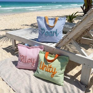May include: Three tote bags on a beach. The blue and white striped bag has "Pearl" embroidered in light blue. The pink bag has "Riley" embroidered in a matching color, and the green bag has "Unity" embroidered in orange. All bags have contrasting colored handles.