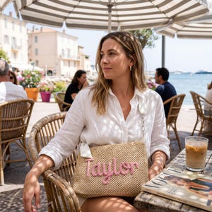 Puede incluir: Un bolso de hombro de paja tejida con el nombre "Taylor" bordado en rosa en cursiva. El bolso se lleva sobre una camisa blanca. Un vaso de caf&eacute; helado y una revista est&aacute;n sobre la mesa. La escena es al aire libre bajo una sombrilla a rayas.