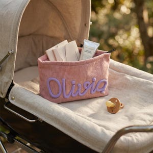 May include: A pink corduroy storage basket with the name "Olivia" in lavender script, filled with small packages and a tube of hand cream. A wooden pacifier rests on the stroller's neutral-toned interior.