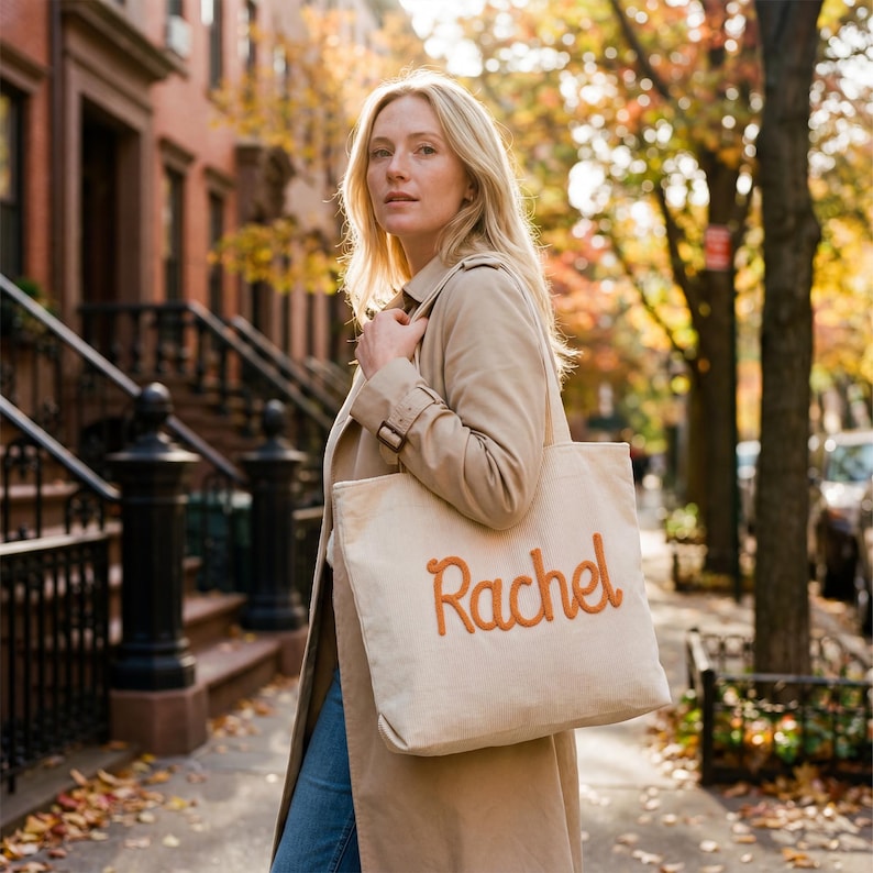 May include: A beige canvas tote bag featuring the name Rachel embroidered in a raised, burnt orange cursive font. The bag has two shoulder straps and a rectangular shape, constructed from a sturdy fabric material with visible stitching details.