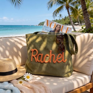 May include: An olive green corduroy tote bag featuring the name Rachel embroidered in large orange script lettering on the front. A pair of dark-rimmed sunglasses rests on the bag, and a fashion magazine is tucked inside the main compartment.
