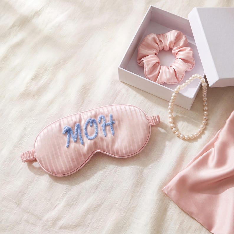 May include: A light pink striped sleep mask with the embroidered blue text MOH, a matching pink scrunchie inside a white gift box, and a single strand of white faux pearls. The items are arranged together on a soft, light-colored fabric surface.
