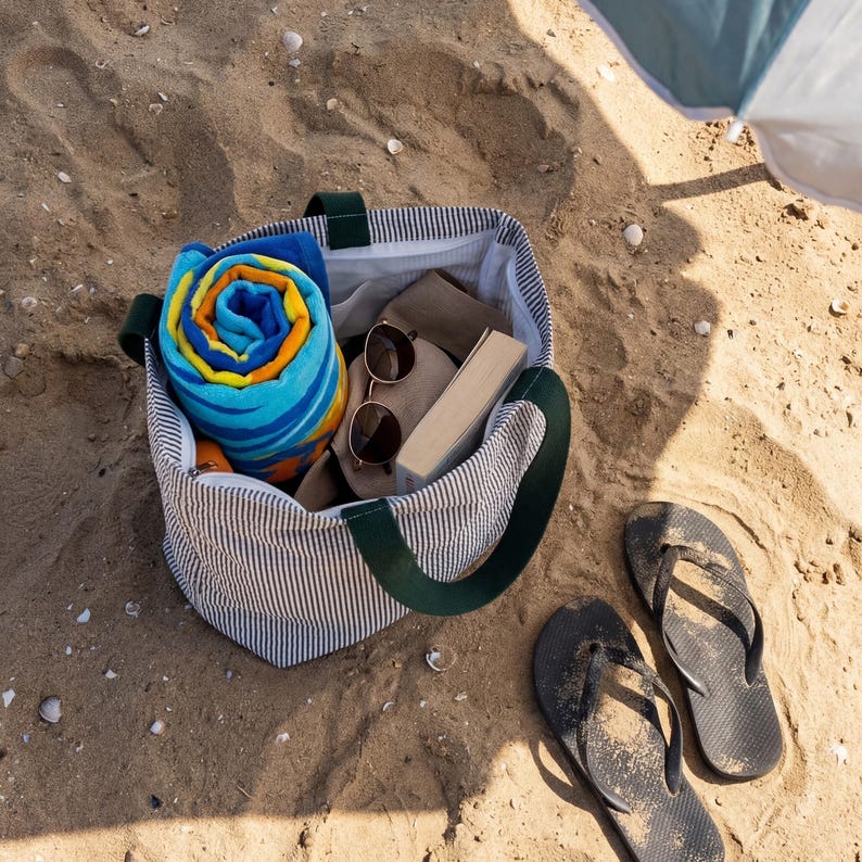 May include: A striped tote bag with green handles, filled with a rolled blue and orange beach towel, sunglasses, a book, and a hat. Black flip-flops rest on the sand next to the bag, suggesting a day at the beach.